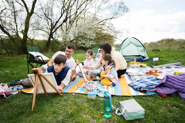 Feliz familia joven con cuatro niños divirtiéndose y disfrutando al aire libre en una manta de picnic pintando en el parque de primavera del jardín relajación