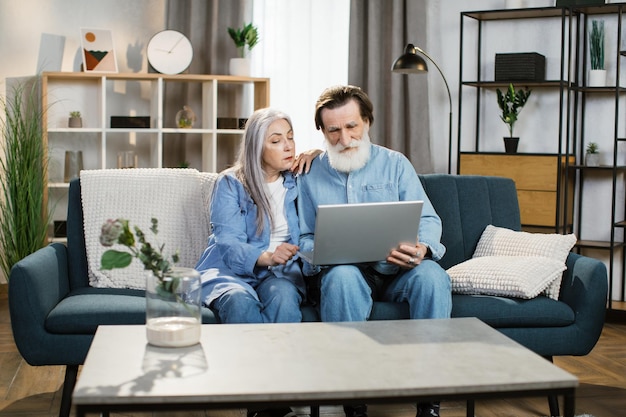 Feliz familia madura con ropa informal sentada en el sofá y usando una laptop inalámbrica