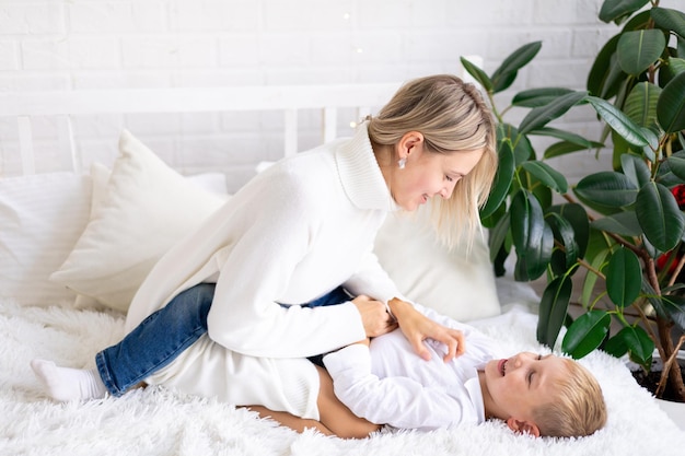 Feliz família mãe e filho em suéteres brancos e jeans na cama em um quarto brilhante em casa abraçando e sorrindo