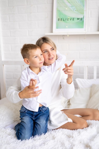 Feliz família mãe e filho em suéteres brancos e jeans na cama em um quarto brilhante em casa abraçando e sorrindo