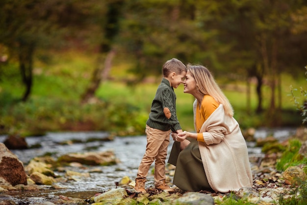 Feliz família mãe e filho, vestindo roupas de malha na margem de um rio de montanha no outono em uma caminhada com ternura se olham.