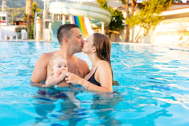 Feliz família mãe pai e filha bebê estão nadando na piscina com toboáguas e se divertindo nas férias beijando e abraçando