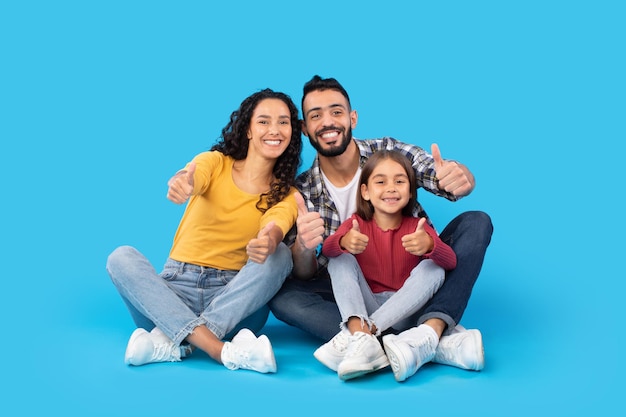 Foto feliz familia del medio oriente gesticulando con los pulgares hacia arriba sobre el fondo azul.