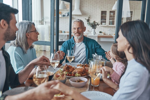 Feliz familia multigeneracional tomados de la mano y rezando mientras cenan juntos