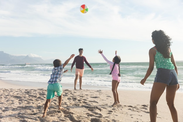 Feliz familia multirracial jugando con pelota en la arena de la playa contra el cielo durante el día soleado