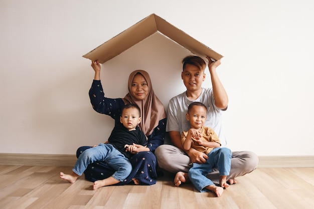 Feliz familia musulmana asiática sosteniendo un cartón que forma el techo de una casa mientras está sentado en el suelo en casa