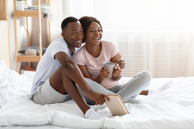 Feliz familia negra que se queda en casa el fin de semana, sentada en la cama, bebiendo té y leyendo un libro, mirando el espacio de la copia. Alegre pareja afroamericana disfrutando de tiempo juntos, pasar el día en la cama