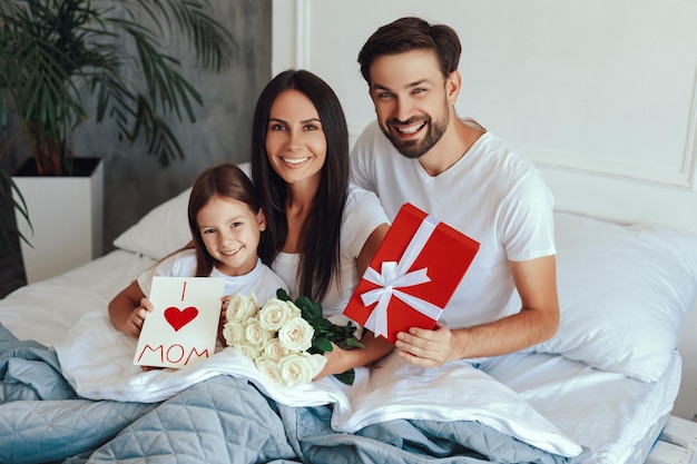 Feliz familia pequeña sonriendo y quedándose debajo de las sábanas mientras sostiene los regalos del día de la madre y mira la cámara