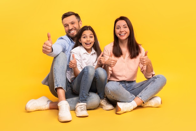 Foto feliz familia de tres dando pulgares hacia arriba sentado con telón de fondo amarillo