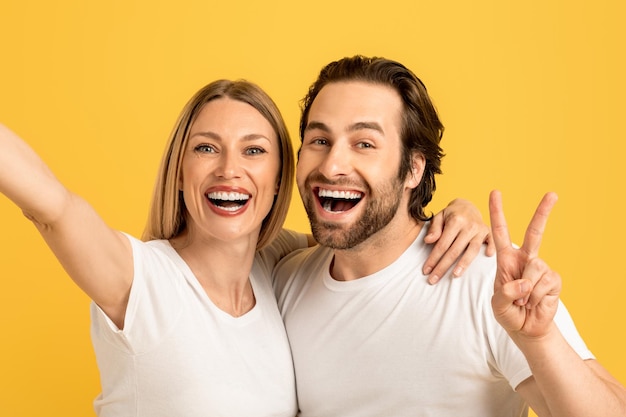 Feliz feliz marido e esposa caucasianos do milênio em camisetas brancas se divertem fazendo sinal de paz e selfie