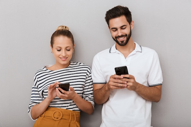 feliz feliz pareja amorosa positiva jóvenes amigos hombre y mujer posando en el interior aislado sobre la pared gris mediante teléfonos móviles.