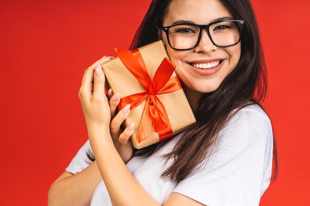 Feliz feriado Retrato de estúdio de uma jovem linda morena sorridente segura caixa de presente isolada sobre fundo vermelho