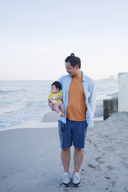 Feliz férias em família na Ásia, um pai segura seu bebezinho na praia no verão, ele olhando para seu bebê, viagem marítima em família