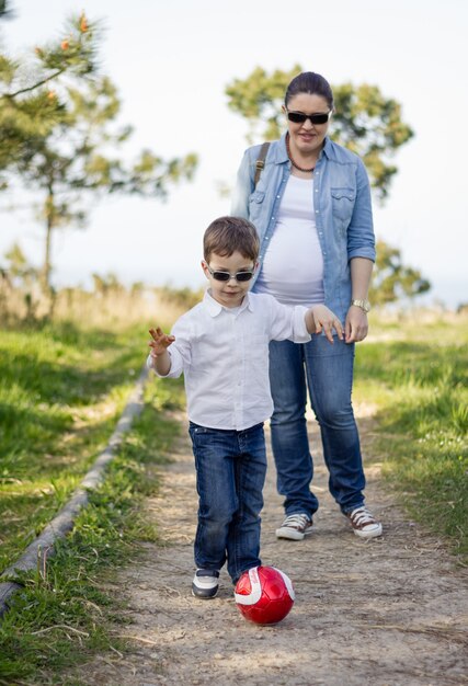 Feliz filho fofo brincando com uma bola de futebol no parque e sua mãe grávida olhando para o fundo