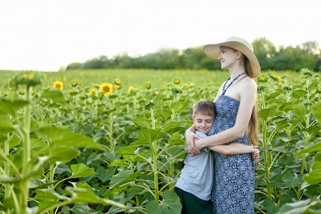 Feliz filho pequeno abraça a mãe grávida em pé em um campo de girassóis florescendo