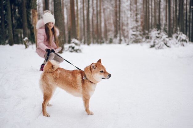 Feliz fin de semana familiar - niña linda en ropa rosa cálido caminar divirtiéndose con el perro rojo shiba inu en el bosque de invierno frío blanco como la nieve al aire libre