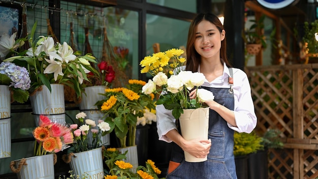 Feliz florista asiática segurando um vaso de flores rosas em frente à sua loja