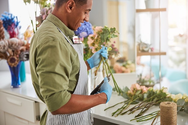 Feliz florista guapo está usando tijeras para cortar tallos de flores antes de venderlos a los clientes