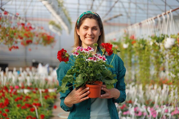 Foto feliz florista mulher bonita com avental trabalhando na estufa enquanto segurando o vaso de flores vermelho e