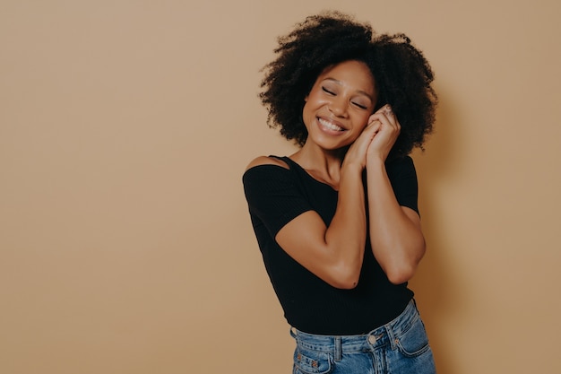 Feliz fofa mulher africana dos anos 20 com cabelo encaracolado, mantendo as mãos perto do rosto e olhos fechados, sorrindo agradavelmente enquanto sonha com algo bom, isolado sobre fundo bege. conceito de emoções humanas