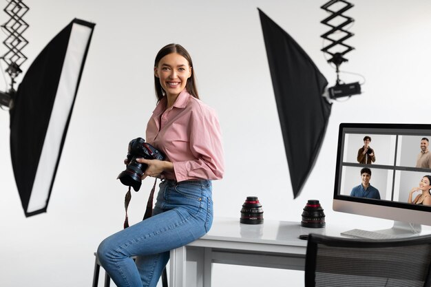 Foto feliz fotógrafa creativa sentada en el lugar de trabajo de la mesa sosteniendo su cámara réflex digital trabajando en