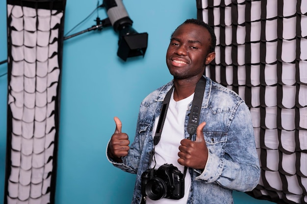 Feliz fotógrafo mostrando o gesto de aprovação em pé no estúdio de produção equipado com conjunto de iluminação profissional e softboxes. Produtor com câmera DSLR posando em fundo azul.