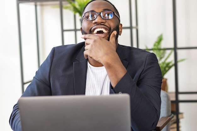 Feliz freelancer jovem afro-americano bem sucedido ou gerente sentado em uma mesa no escritório; regozijando-se com um negócio bem-sucedido ou novo projeto de negócios