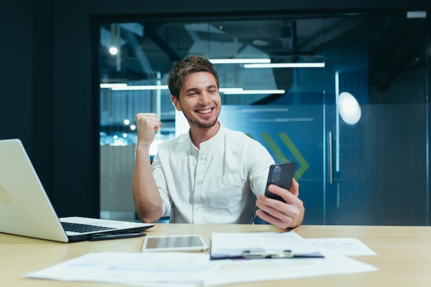 Feliz freelancer trabalha no escritório com laptop sorri e olha para a tela do telefone se alegra com o gesto de vitória do sucesso