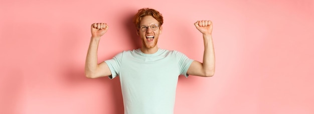 Feliz ganador con pelo rojo y barba celebrando la victoria gritando sí con alegría y levantando las manos