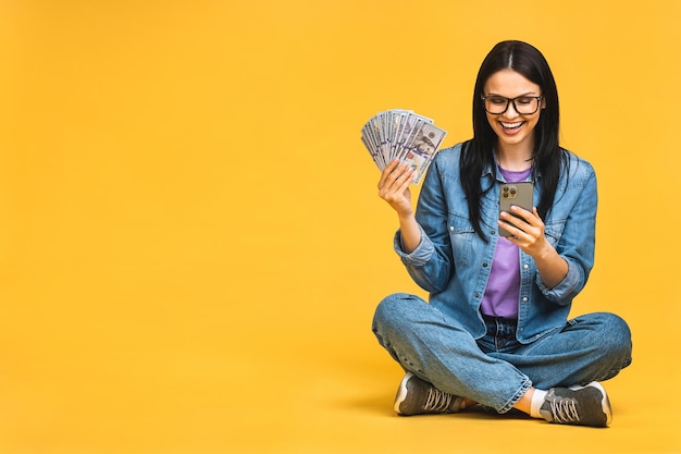 Foto feliz ganador retrato de una mujer alegre y sonriente usando un smartphone aislado sobre un fondo amarillo sentada en el suelo en posición de loto sosteniendo billetes de dinero