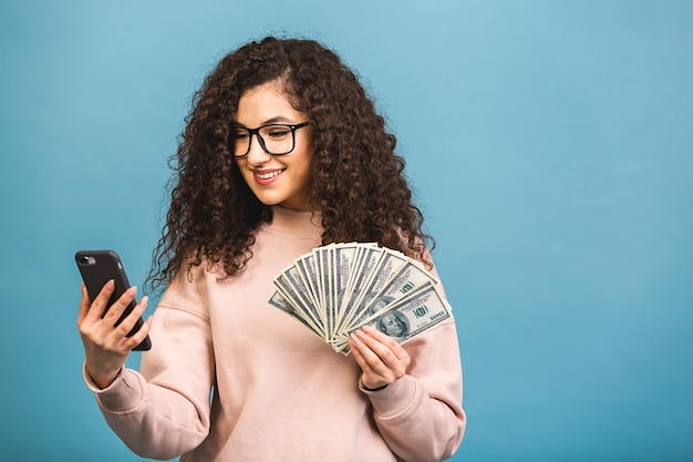 ¡Feliz ganador! Retrato de una mujer rizada joven alegre sosteniendo billetes de banco y celebrando aislado sobre fondo azul. Usando el teléfono.