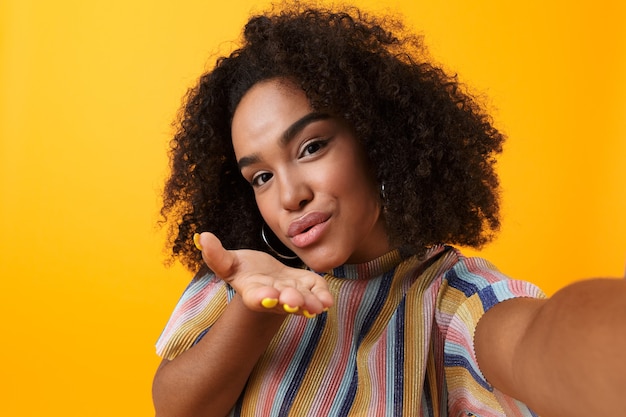 Feliz garota africana bonita posando isolado sobre o espaço amarelo tomar uma selfie mandando beijos.