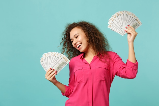 Foto feliz garota africana em roupas casuais, segurando um leque de dinheiro nas notas de dólar, dinheiro isolado no fundo da parede azul turquesa. emoções sinceras de pessoas, conceito de estilo de vida. simule o espaço da cópia.