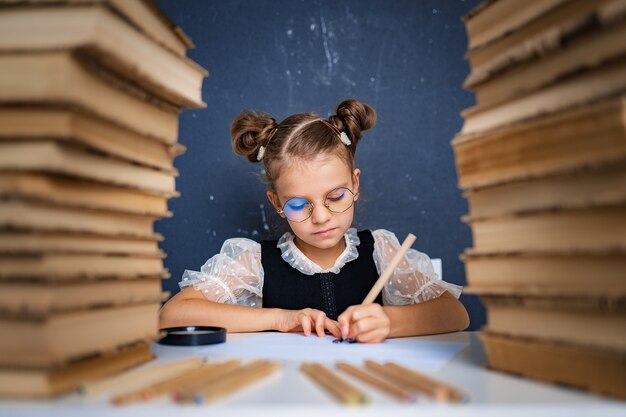 Feliz garota esperta de óculos arredondados, escrevendo, desenhando enquanto está sentado entre duas pilhas de livros.