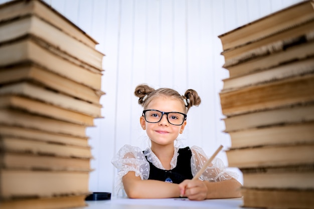 Feliz garota esperta de óculos arredondados, segurando um lápis na mão pronto para escrever sentado entre duas pilhas de livros e olhar para a câmera sorrindo.