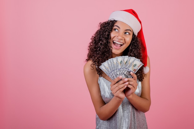 Feliz garota negra segurando notas de cem dólares, usando chapéu de Papai Noel isolado