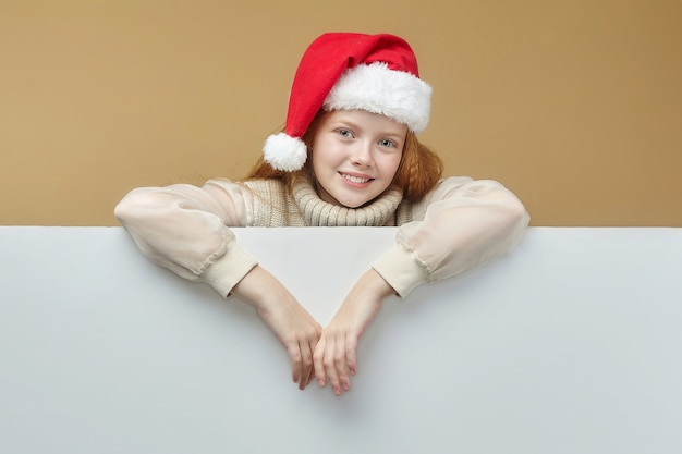 Feliz garota ruiva com um chapéu de papai noel e um outdoor branco