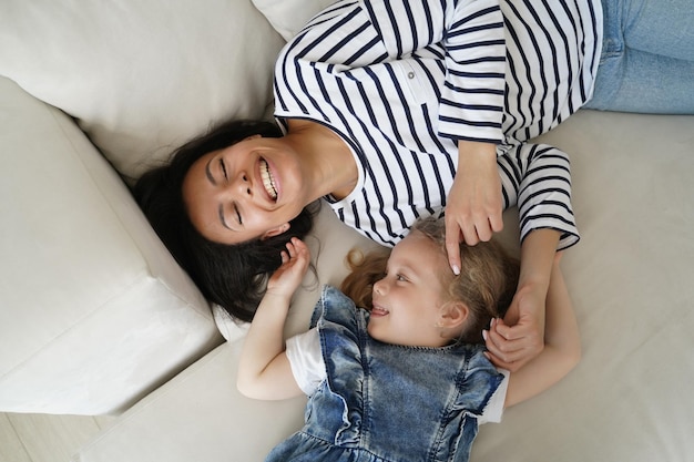 Feliz garotinha adotada fazendo cócegas na mãe rindo deitada se divertindo juntos na vista superior do sofá