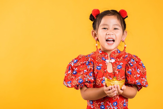 Feliz garotinha chinesa asiática sorrindo usando cheongsam vermelho segurando lingote de ouro