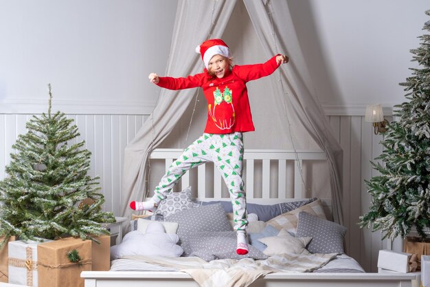 Feliz y graciosa niña en pijama y gorro de Papá Noel salta en la cama en la mañana de Navidad El niño se está divirtiendo