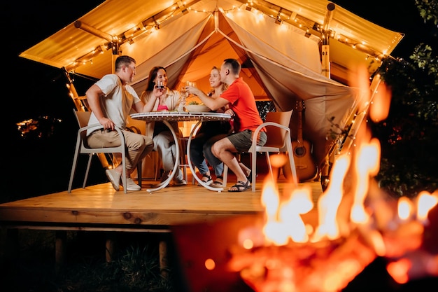 Feliz grupo de amigos relaxando em glamping e bebendo vinho na noite de verão perto da fogueira aconchegante Barraca de acampamento de luxo para recreação ao ar livre e recreação Conceito de estilo de vida