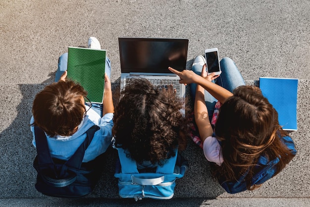 Feliz grupo de escolares preadolescentes sentados y usando dispositivos digitales y portátiles al aire libre