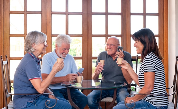 Feliz grupo de personas mayores caucásicas sentadas en la mesa disfrutando de una taza de café y usando el teléfono