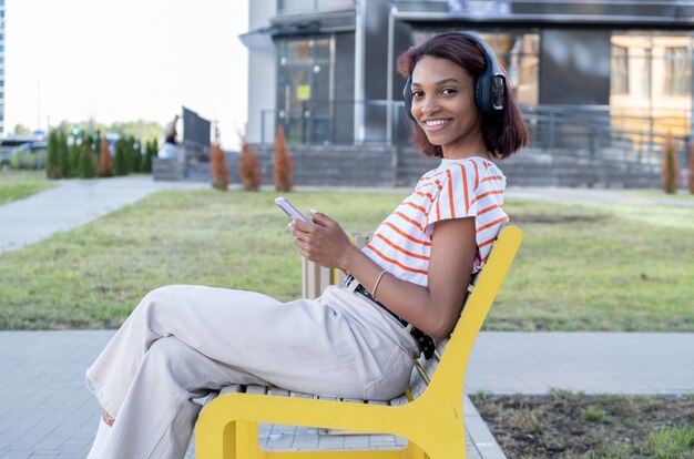 Feliz guapa joven morena disfrutando de su música con auriculares estéreo sentada contra una puerta enrollable de madera con su teléfono móvil