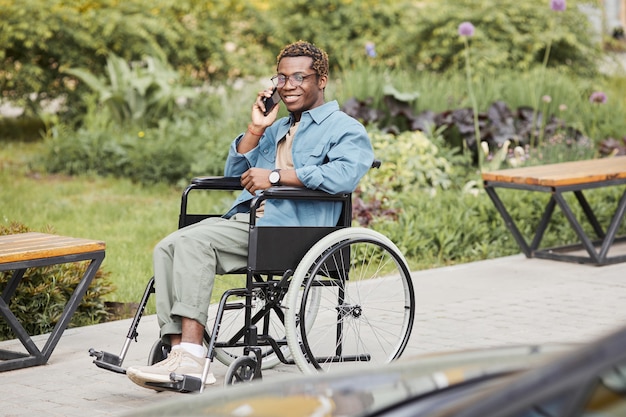 Feliz guapo joven afroamericano discapacitado en anteojos sentado en silla de ruedas y hablando por teléfono