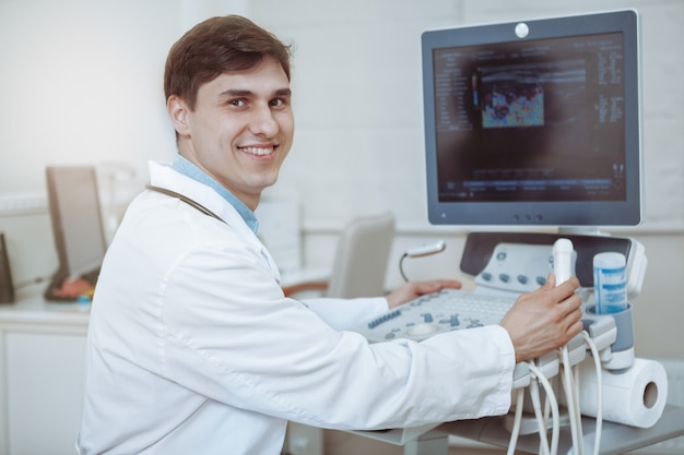 Feliz guapo médico masculino sonriendo a la cámara, trabajando en la máquina de ultrasonido en su clínica