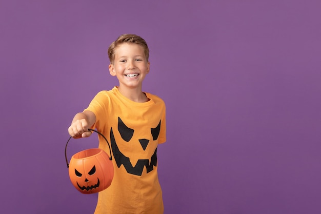 Feliz Halloween Sonriente niño preadolescente en camiseta de Halloween sosteniendo busket de calabaza permaneciendo sobre fondo púrpura Disparo de estudio