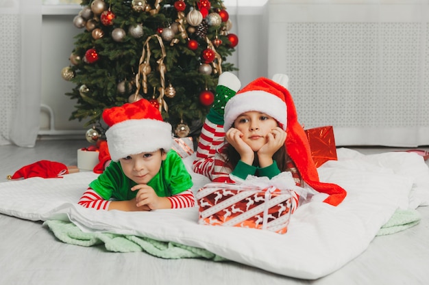 Feliz hermano y hermana vestidos como Santa Claus están sentados cerca del árbol de Navidad en casa en la sala de estar con regalos. Navidad