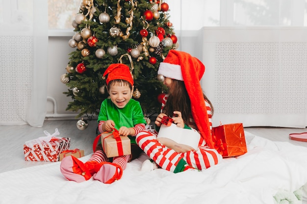 Feliz hermano y hermana vestidos como Santa Claus están sentados cerca del árbol de Navidad en casa en la sala de estar con regalos. Navidad