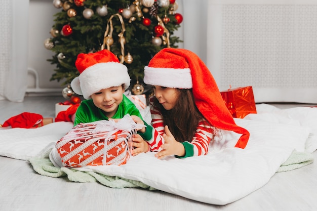 Feliz hermano y hermana vestidos como Santa Claus están sentados cerca del árbol de Navidad en casa en la sala de estar con regalos. Navidad